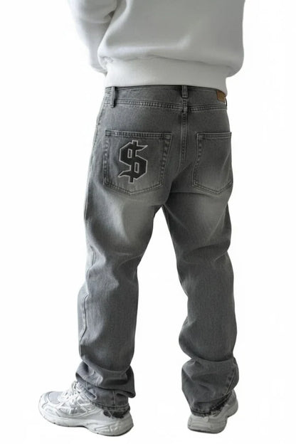 A person shown from the back wearing gray denim jeans with a large black dollar sign patch on the back pocket, paired with a white sweatshirt and white sneakers, against a plain white background.