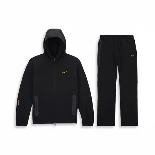 A Black Nike nocta athletic, loose fit tracksuit set consisting of a zip-up hooded jacket and matching jogger pants, both featuring a small white Nike swoosh logo on the chest, displayed against a solid white background.